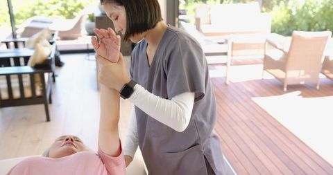 Physical therapist assisting senior woman with arm stretch on table in sunlit clinic room