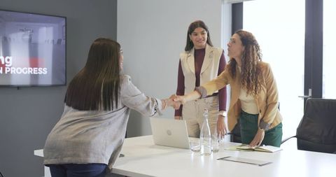 Businesswomen shaking hands in modern boardroom during meeting with laptop presentation