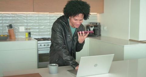 Remote working man speaking into phone at modern kitchen island with laptop and coffee mug