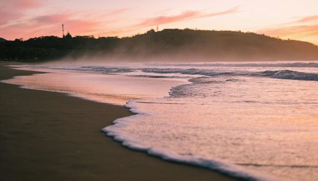 Pastel sunset beach with curving foam line, reflective wet sand and misty headland