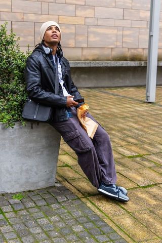 Young man sitting on planter edge eating sandwich and checking phone in urban plaza