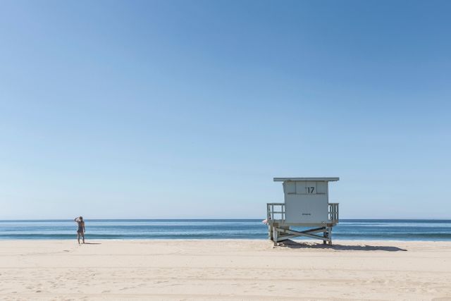 Woman stands on pristine sandy beach near lifeguard stand under clear blue sky. Ideal for summer vacation promotions, travel guides, beach lifestyle features, and tranquil getaway advertisements.