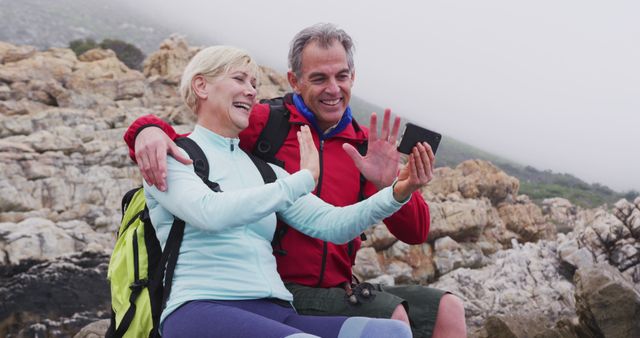 Senior hiker couple with backpacks sitting on the rocks and waving while having a image call on smartphone while hiking near sea shore. trekking, hiking, nature, activity, exploration