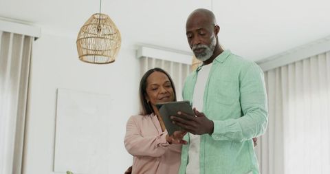 Mature black couple standing at home using tablet together under rattan pendant lamp