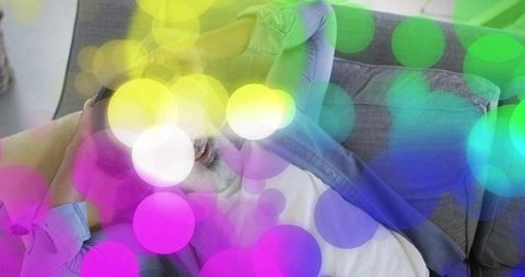 Senior man reclining, laughing and relaxing on grey sofa with colorful bokeh overlay