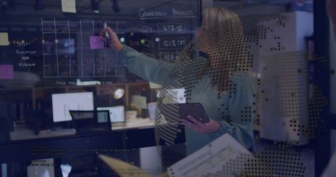 Businesswoman marking glass grid holding tablet in modern office with data overlay