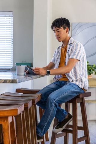Asian man sitting at kitchen island checking smartphone while holding coffee mug