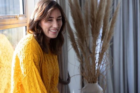 Woman smiling by sunlit window wearing mustard knit sweater with cozy pampas grass