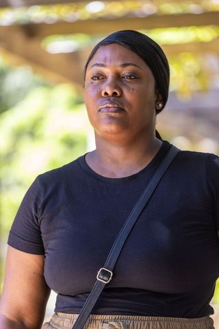 Contemplative woman standing under sunlit pergola, wearing black tee and crossbody strap