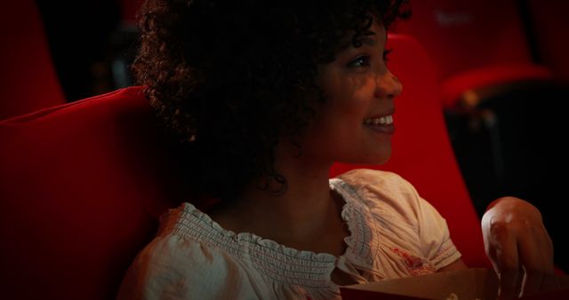 Young woman with curly hair enjoying popcorn while watching a movie in a theater. Brightly lit face with a smile. Great for illustrating themes of relaxation, entertainment, leisure activities, and enjoying personal time.