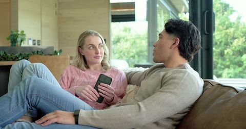 Couple Relaxing on Sofa Talking and Holding Smartphone in Modern Daylit Living Room