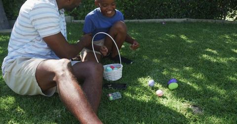 African american father and son hunting easter eggs sunlit backyard lawn smiling together