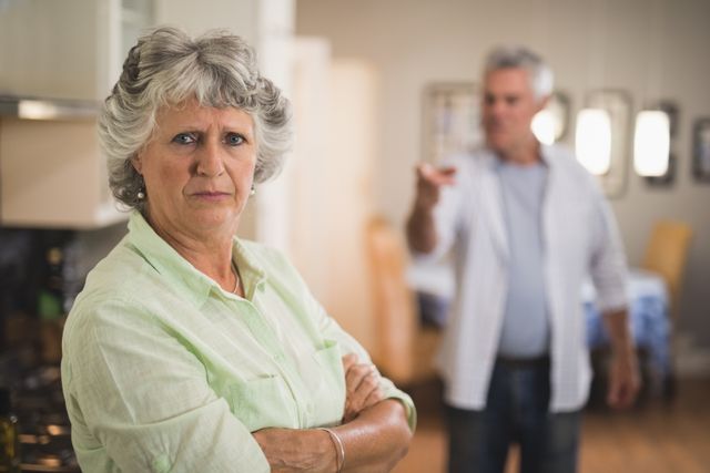 Senior woman stands with a serious expression and crossed arms while an angry man gestures in the background. Ideal for illustrating themes of relationship issues, family dynamics, and domestic conflict. Useful for articles, blogs, and advertisements focusing on elderly relationships, conflict resolution, and emotional well-being.