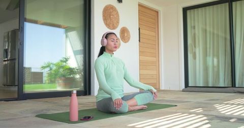 Middle-aged asian woman meditating on sunlit patio wearing mint top and grey leggings