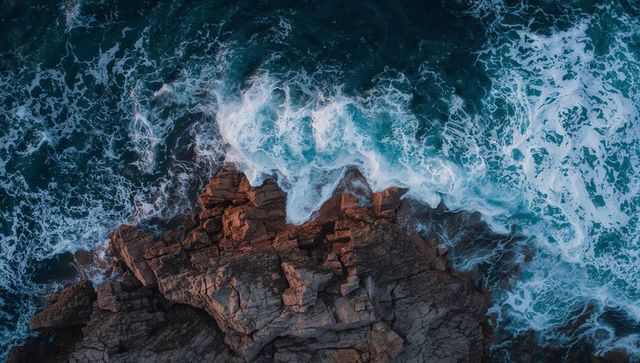 Waves crashing against jagged coastal rock from aerial view with teal and navy foamy surf