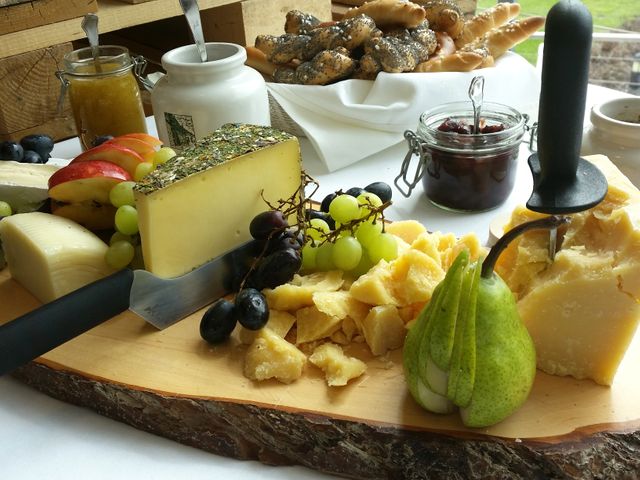 Cheese platter with an assortment of cheeses, fresh fruits, and a knife on a rustic wooden board, presented outdoors. Grapes, sliced apples, and a pear complement the setup. Ideal for showcasing gourmet appetizers, outdoor dining experiences, and rustic food presentations.
