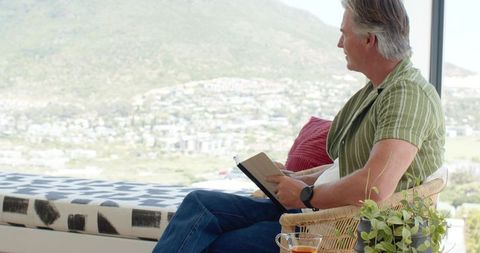 Mature man reading by panoramic window overlooking sunlit valley in cozy wicker chair