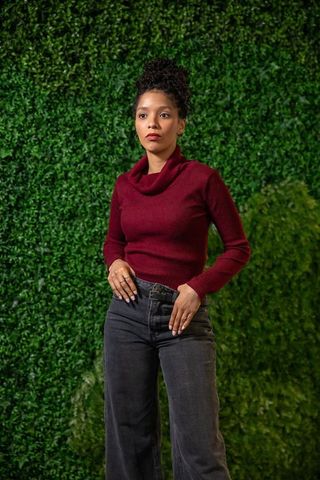 Confident african american woman wearing burgundy sweater standing by green foliage wall