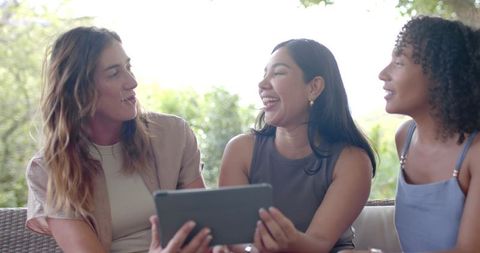 Diverse women laughing and sharing tablet on sunlit patio sofa, casual summer friends