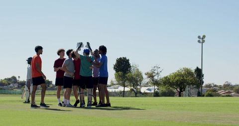 Youth cricket team celebrating in huddle on sunny grassy pitch wearing colorful tees