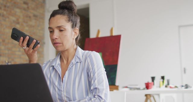 An ambitious businesswoman is working diligently on her laptop while using her smartphone for a business call. She is sitting in a modern office space with an easel in the background, indicating a creative environment. This image can be used in articles or posts about remote work, multitasking, business communications, and modern workspaces.