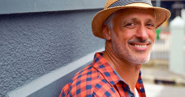 Mature man with a happy expression wearing a straw hat and plaid shirt, leaning on a textured wall outdoors. Great for use in lifestyle, summer, men's fashion, confidence, and outdoor activity concepts.