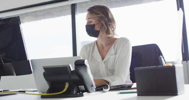 Businesswoman wearing face mask sitting at desk with computer in modern office, reflecting health and safety protocols during COVID-19 pandemic. Suitable for articles, advertisements, or resources related to workplace safety, COVID-19 measures, professional environments, and office work.