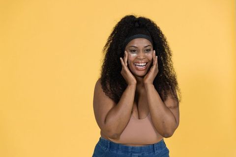 Confident plus-size african american woman smiling applying eye cream, casual jeans, yellow