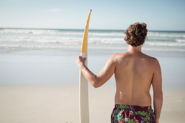 Rear view of shirtless man holding surfboard at beach during sunny day