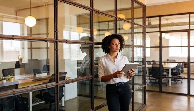 Professional woman standing holding tablet by industrial glass wall reflecting modern office