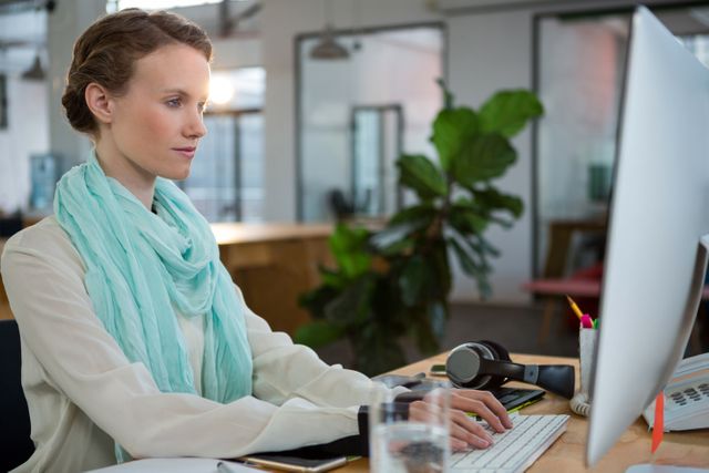 This image depicts a female graphic designer working intently at her desk in a contemporary office environment. The setting is bright and modern, featuring ergonomic office furniture and plants that add a touch of nature. This can be used for articles or websites related to graphic design, female professionals in creative fields, modern office design, or productivity tips.