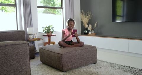 African teen girl sitting cross-legged on ottoman wearing headphones using phone in living room