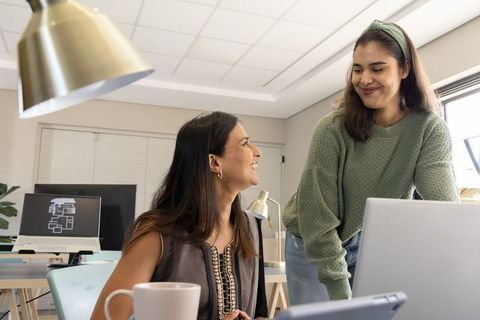 Diverse female coworkers collaborating at desk with laptop and floorplan on monitor