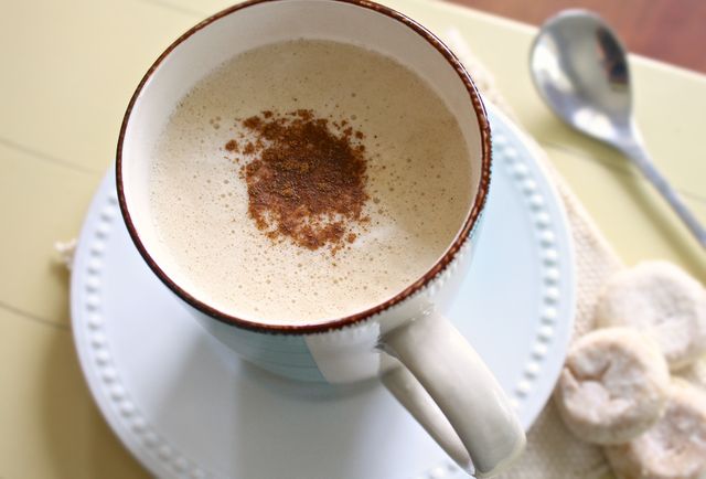 Close-up image showing a steaming, frothy cappuccino with a sprinkle of cinnamon on top. Ceramic mug is placed on a light blue saucer with a spoon beside it. Ideal for use in content related to coffee culture, breakfast routines, cozy autumn or winter themes, and beverage advertising.