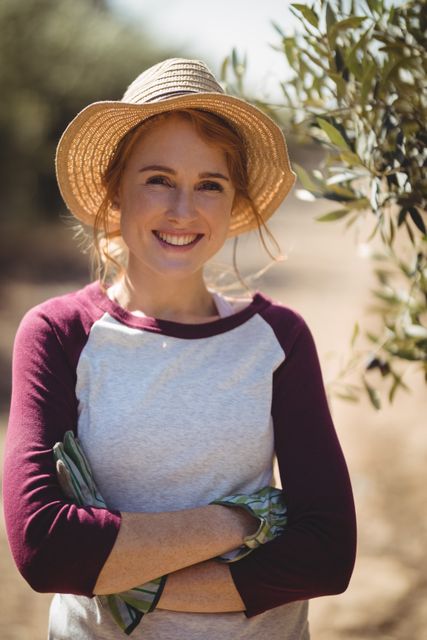 This image shows a young woman smiling and standing confidently at an olive farm. She is wearing a straw hat and casual clothing, with her arms crossed. This image can be used for articles or advertisements related to agriculture, farming, rural life, or outdoor activities. It is also suitable for promoting agricultural products or services, as well as for lifestyle blogs focusing on countryside living.