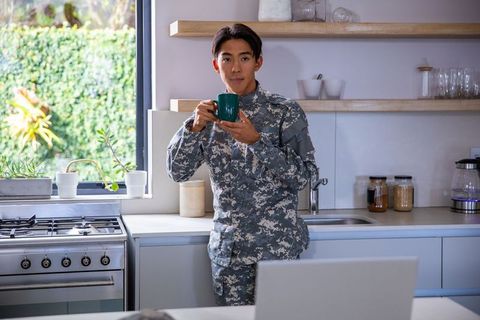Asian soldier standing in modern kitchen holding green mug near laptop, morning coffee