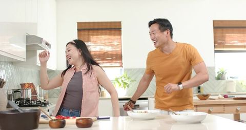 Asian couple cooking and laughing at bright modern kitchen island, chopping fresh veggies