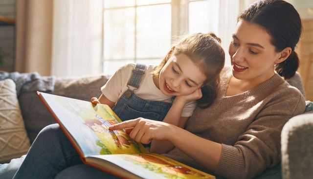 Mother and daughter reading together on cozy sofa by sunny window - family bonding