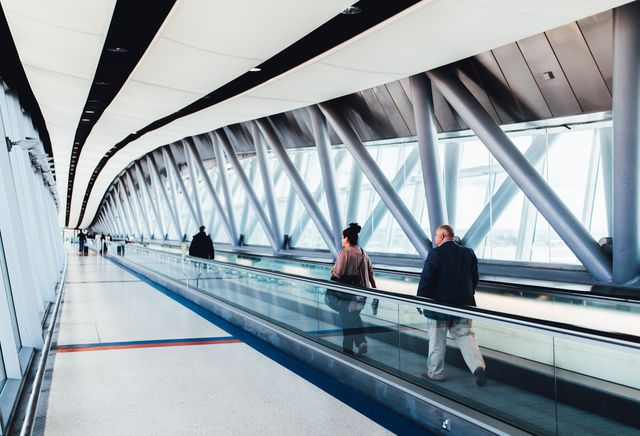 Business and leisure travelers walking on indoor airport moving walkway. Modern architecture with large windows. Useful for articles about air travel, airport amenities, transportation infrastructure, and business commuting.