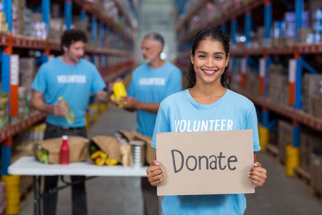 Young woman holding a 'Donate' sign in a warehouse, smiling at the camera. Other volunteers are seen in the background sorting items. Ideal for use in campaigns promoting charity, community service, and humanitarian efforts. Perfect for illustrating concepts of giving back, teamwork, and social responsibility.