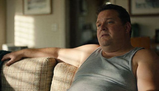Middle-aged man relaxing on sunlit couch in gray tank top, contemplative home leisure