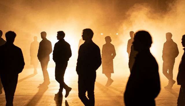 Silhouetted figures standing in amber backlight casting long shadows across foggy plaza