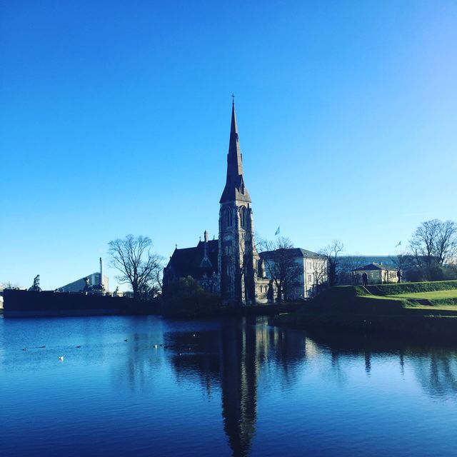 Historic Gothic-style church standing tall by calm lake, reflecting clear blue sky. Perfect for visuals related to travel, historic architecture, serene landscapes, and European tourism. Ideal for website banners, travel brochures, and cultural appreciation content.
