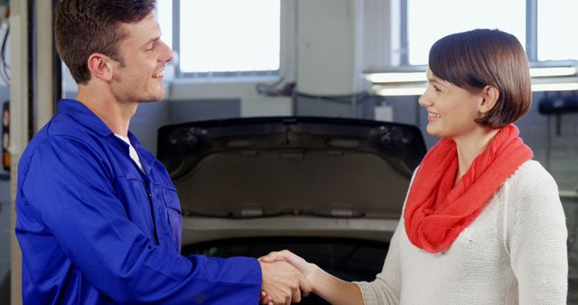 Mechanic wearing blue uniform and smiling woman with red scarf shaking hands in car repair shop. Great for automotive service advertisements, customer service promotions, or business trust-themed projects.