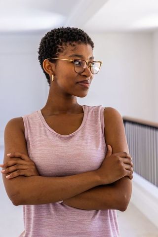 Confident young black woman standing arms crossed wearing gold glasses and pink top
