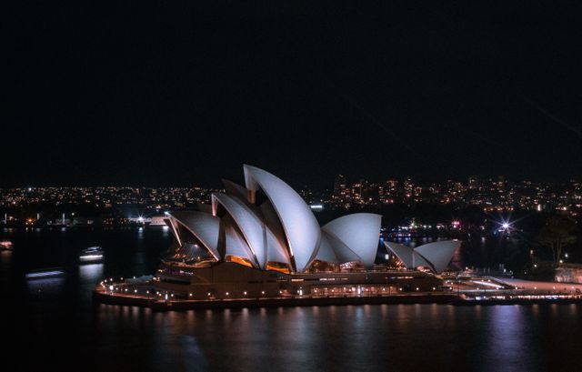 Sydney Opera House glowing against night sky with city lights in background and water reflections. Ideal for travel advertisements, architectural features, cityscape illustrations, tourism promotions, or cultural event posters.