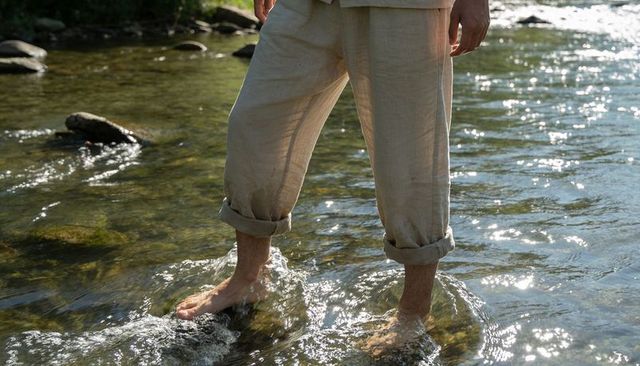 Wading barefoot through sunlit river in rolled linen pants over mossy rocks
