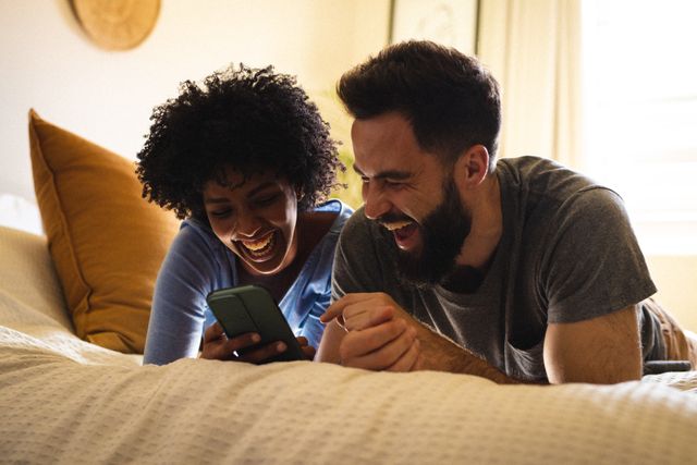 Cheerful biracial young couple using mobile phone and laughing while lying on bed at home. Unaltered, lifestyle, love, togetherness, afro hair, beard, relaxation and technology concept.