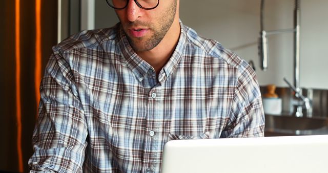 Young professional man wearing glasses and a plaid shirt is working on a laptop in a modern kitchen. Ideal for themes of remote work, home office, casual business environment, and modern lifestyle. Perfect for blogs and articles about work-from-home setups, online business, and professional productivity.