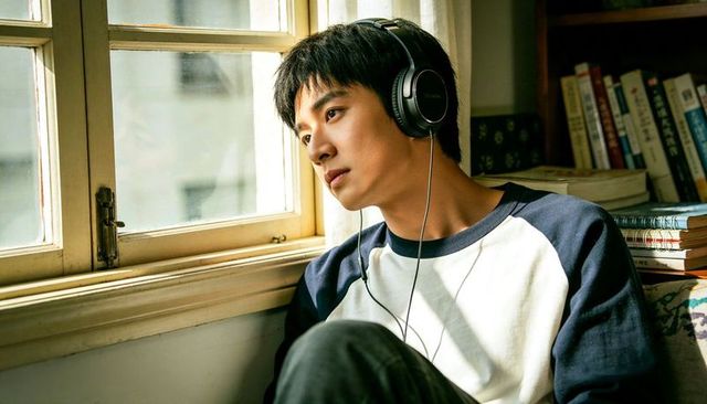 Young man wearing headphones gazing out sunlit window in cozy study, contemplative mood